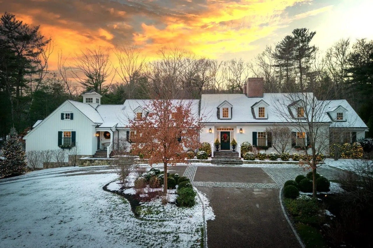 Winter residential landscape with a partially cleared circular driveway, light snow cover, and holiday lighting surrounding a white New England home.
