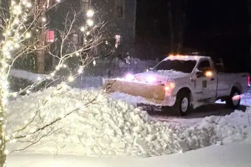 Snowplow clearing a residential driveway at night during a winter storm, with holiday lights illuminating the snowy landscape.