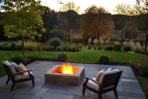 A stone patio and fire pit overlook a lush garden at dusk, creating an inviting outdoor retreat designed for comfortable evenings outside.
