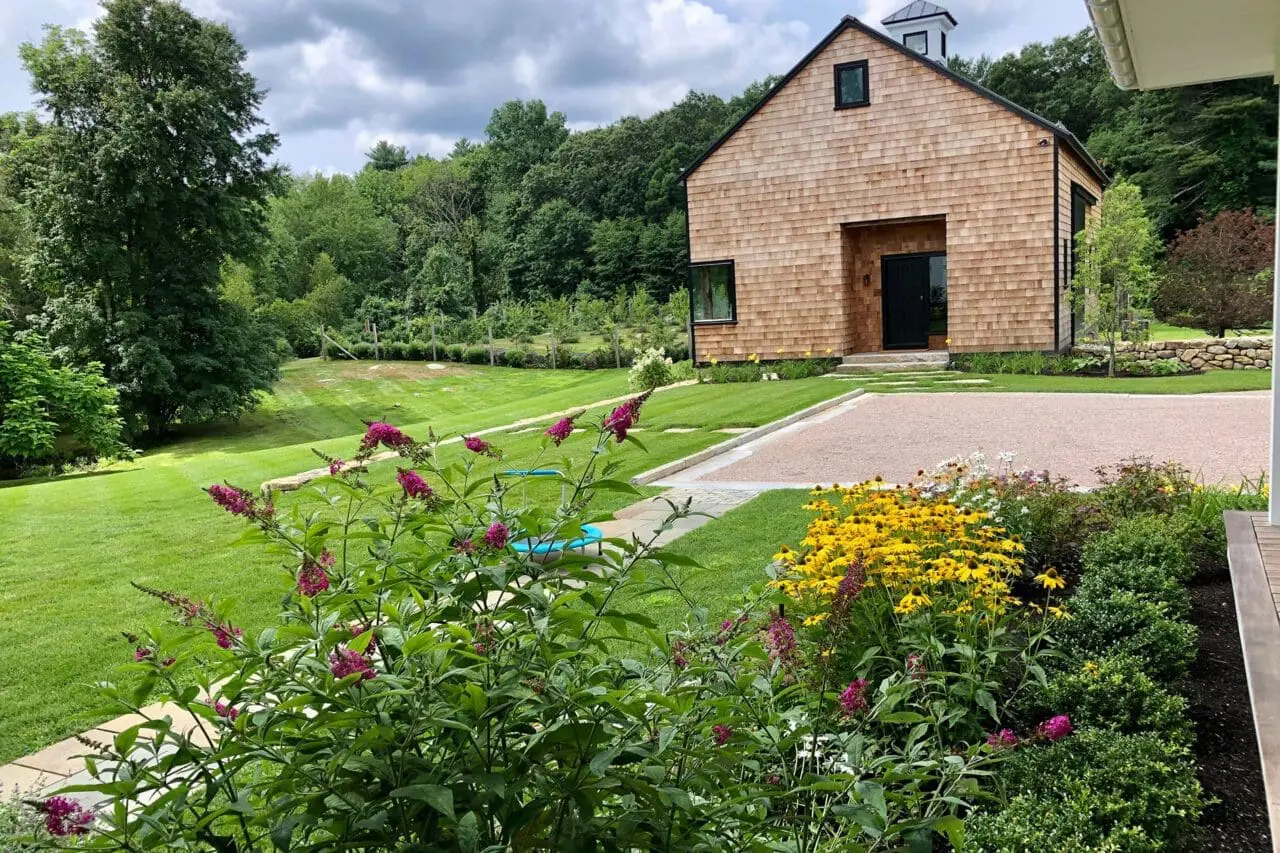 Carlisle, MA - A landscaped garden with native plantings and flowering perennials surrounding a modern cedar-shingled home, blending structure with a natural setting.