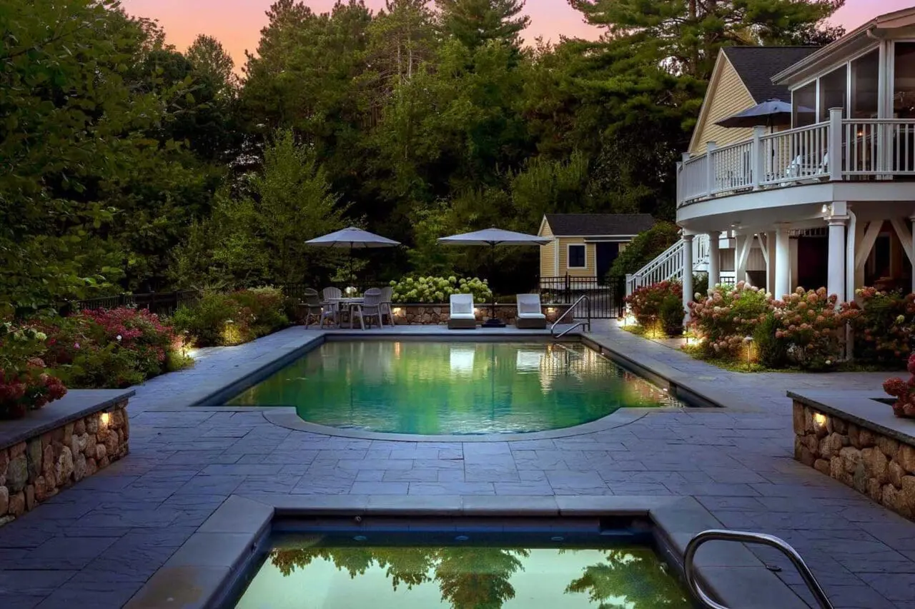 Residential landscape with a swimming pool, spa, stone patio, hydrangeas, outdoor seating, and integrated landscape lighting beside the home.