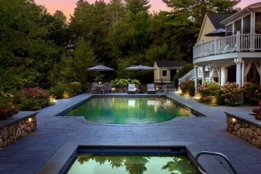 Residential landscape with a swimming pool, spa, stone patio, hydrangeas, outdoor seating, and integrated landscape lighting beside the home.