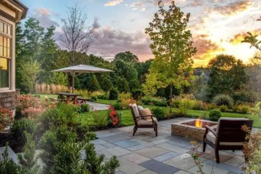 A bluestone patio with a built-in fire feature overlooks lush gardens and layered plantings as the sun sets over this Wayland landscape.
