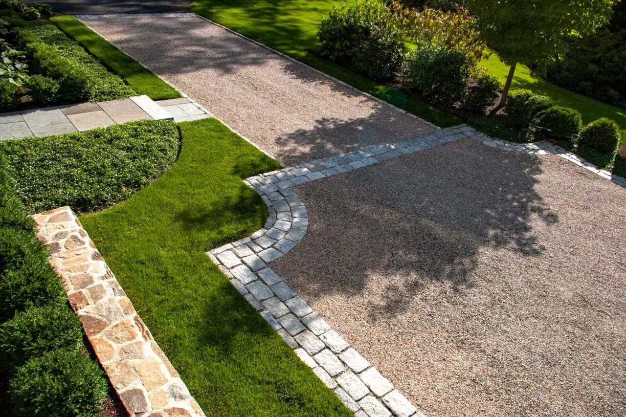 A gravel driveway is shaped and defined by a broad cobblestone border, with crisp lawn edges, low stonework, and neatly maintained plantings creating a polished transition through the property.