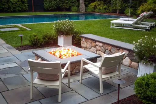 A square stone fire pit anchors this bluestone terrace beside the pool, offering clean lines, durable materials, and a central gathering point that enhances both day and evening use.