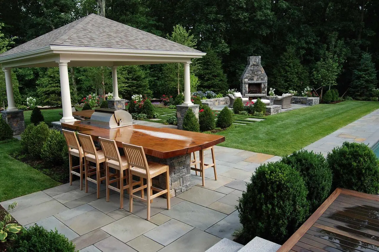 Pavilion-defined dining area on a stone patio pairs a wood table with a built-in grill, adding structure, light shade, and an inviting setting for outdoor entertaining.