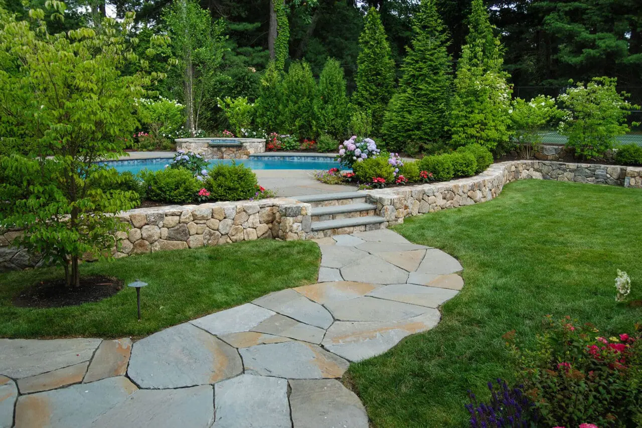 A curved flagstone walkway meets a stone retaining wall near the pool terrace, with shrubs and hedges softening curves and enhancing flow.