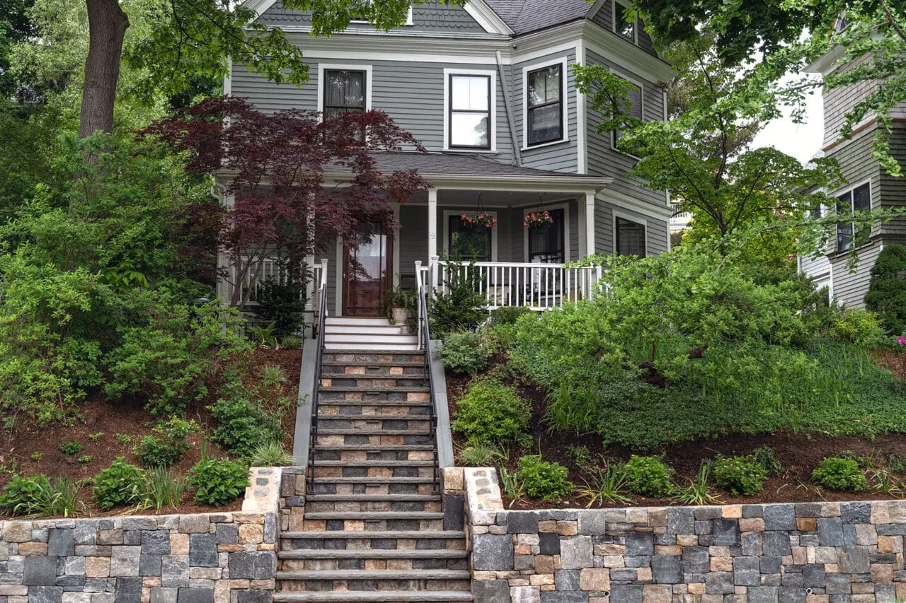 Belmont, MA - Granite entry steps rise from a stone retaining wall in the front yard, using durable materials and surrounding shrubs to create a welcoming, structured approach.