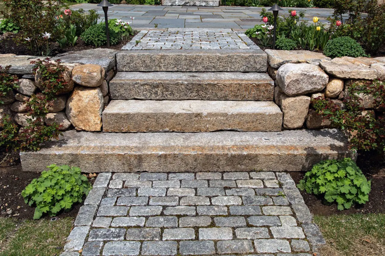 Granite steps descend to a cobblestone landing between fieldstone walls, with shrubs and hedges adding structure and refinement to this entry sequence in Weston.