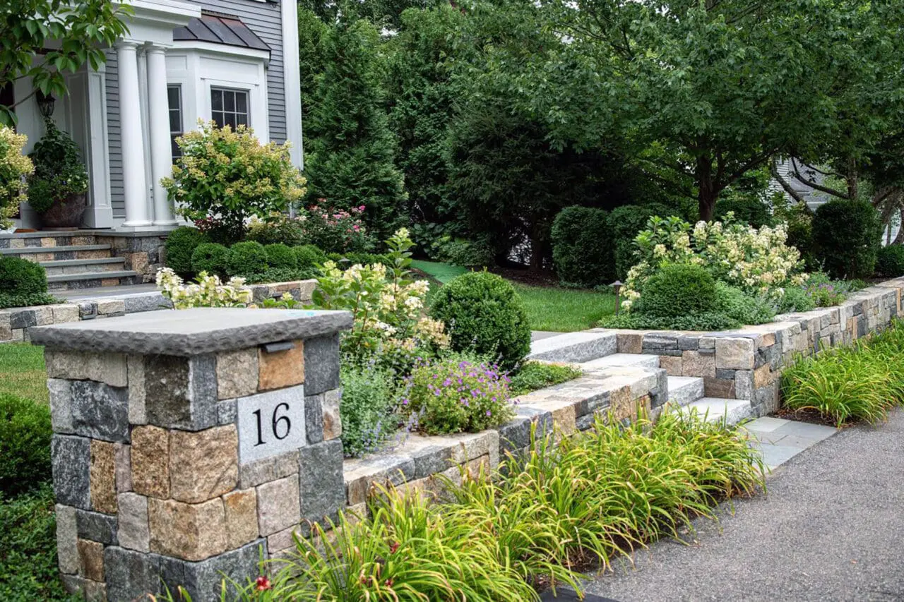 A low stone entry wall paired with granite steps establishes a clear arrival sequence, with adjacent hedges and foundation shrubs adding definition and a welcoming sense of enclosure.