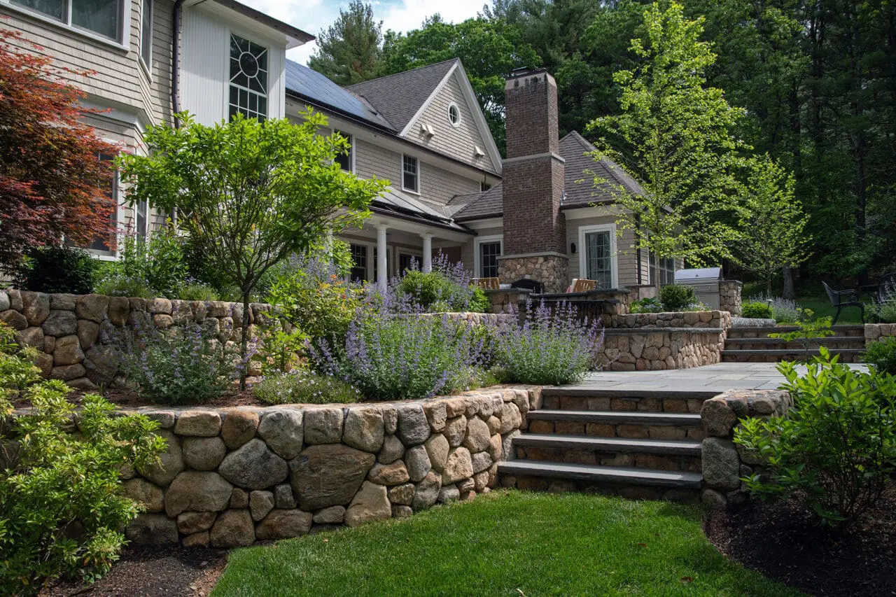 Tiered fieldstone walls paired with bluestone steps create a strong visual connection between the patio and surrounding planting, using layered stonework to manage grade while framing shrubs and low hedges that soften transitions between outdoor rooms.