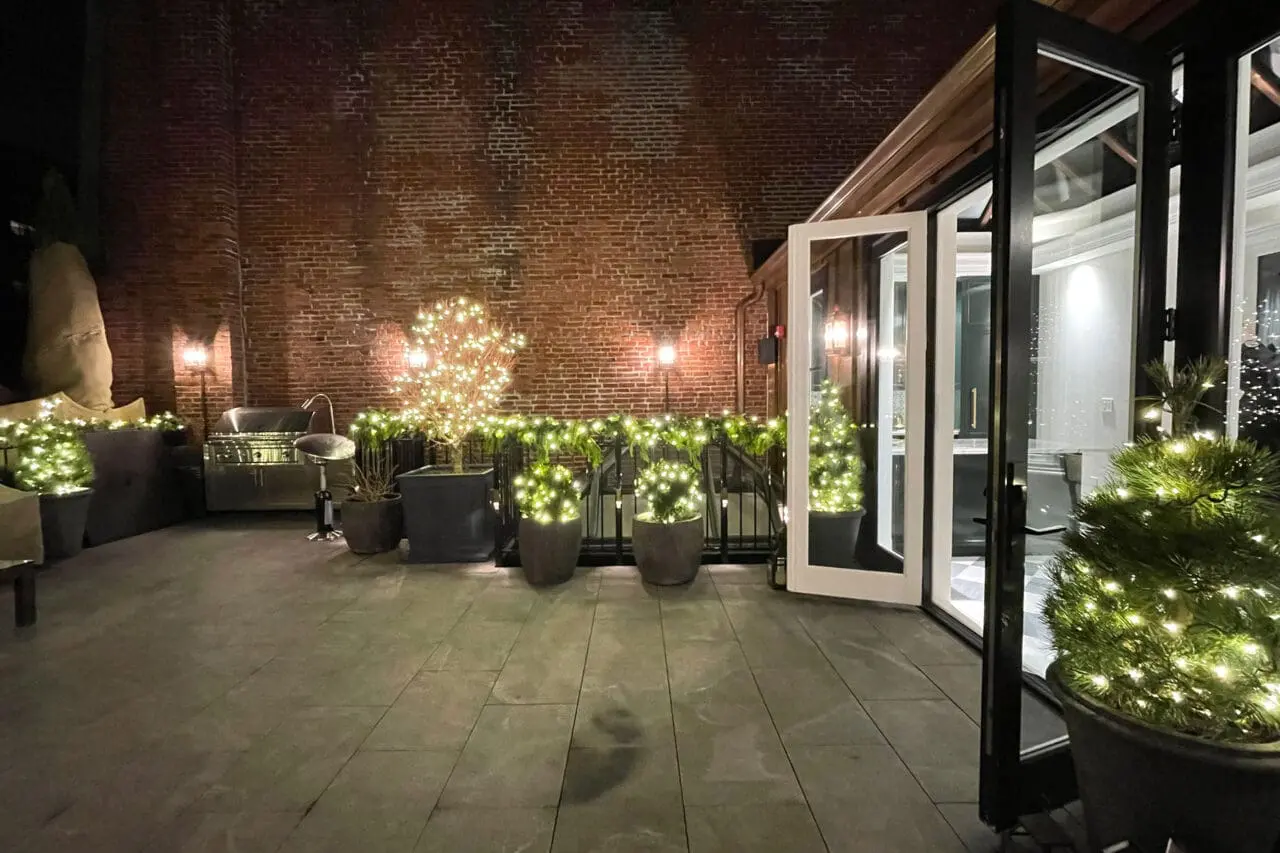 An intimate urban courtyard is dressed for winter with layered evergreen containers, warm string lighting, and seasonal accents that bring a festive glow to the outdoor patio.