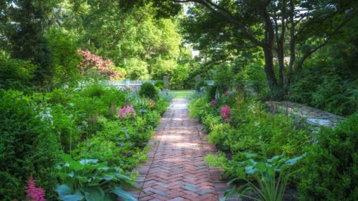A herringbone brick garden path winds through layered perennial plantings beneath mature trees, showing how hardscape and landscaping can work together naturally.