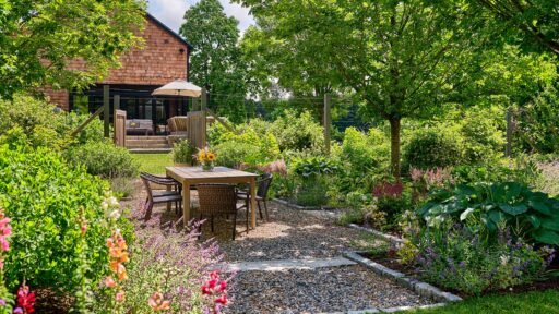 Boston-area garden designed with a gravel dining terrace, layered perennial plantings, mature trees, and a welcoming outdoor seating area.