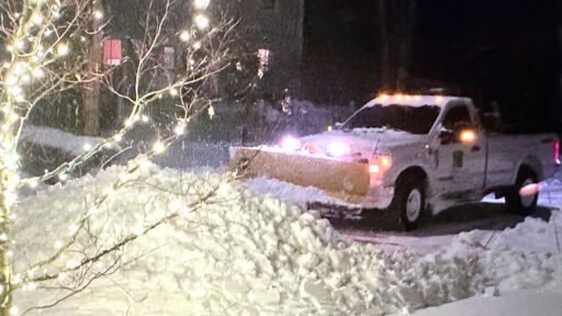 Snowplow clearing a residential driveway at night during a winter storm, with holiday lights illuminating the snowy landscape.
