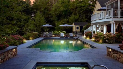 Residential landscape with a swimming pool, spa, stone patio, hydrangeas, outdoor seating, and integrated landscape lighting beside the home.