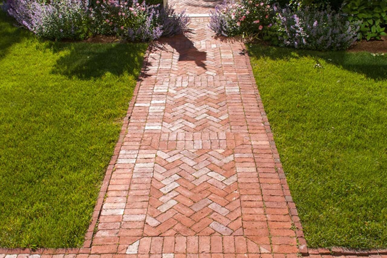 Cambridge, MA - A classic herringbone brick walkway creates a welcoming approach to a historic garden gate, framed by lush perennial plantings and lawn.