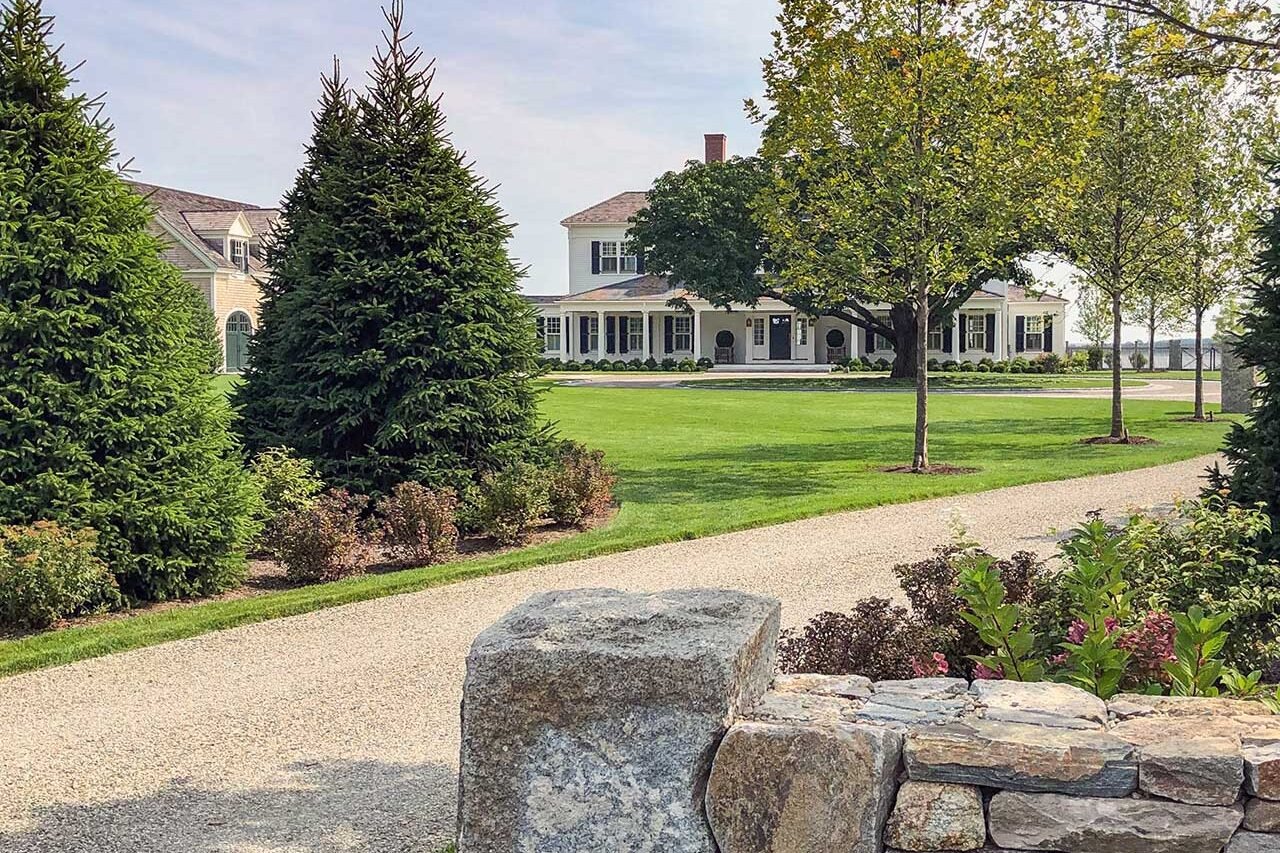 Duxbury, MA - A natural stone wall and gravel drive frame the approach to the home, with mature trees, evergreen plantings, and an expansive lawn creating a welcoming sense of arrival.