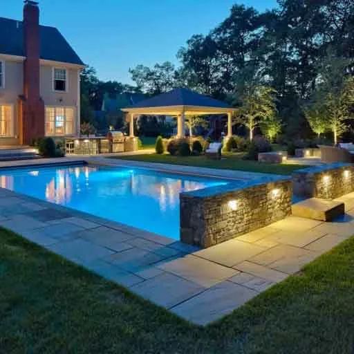 Sudbury, MA - This pool landscape uses stone walls, exterior lighting, and layered outdoor living spaces to create a polished evening setting.