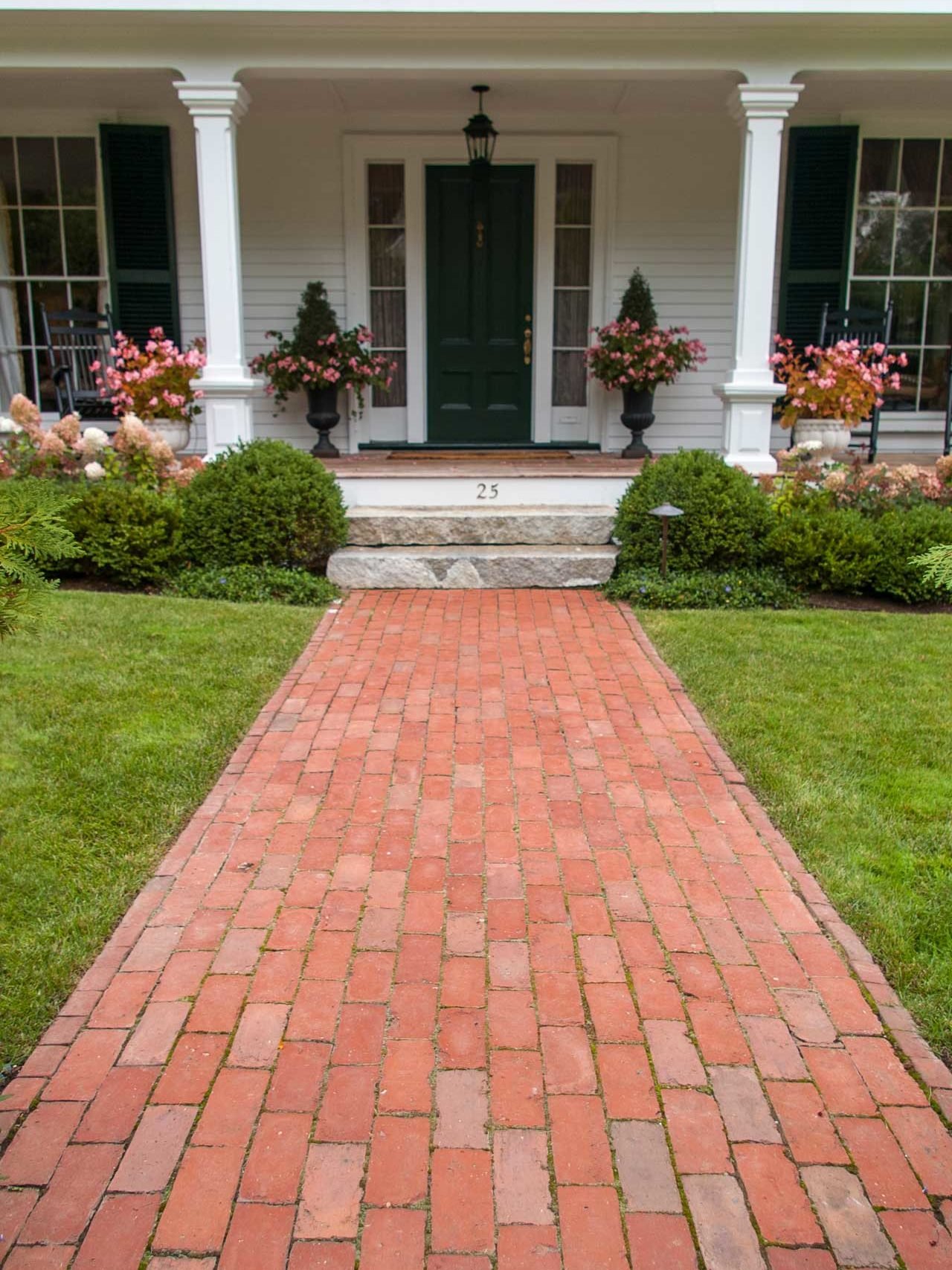 Belmont, MA - Classic clay brick walkway and antique granite steps lead the way to a sprawling front porch accented by colorful planters.