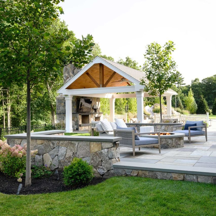 A stone terrace with built-in fire pit and open-air pavilion creates a comfortable outdoor living space framed by lawn, trees, and flowering plantings.