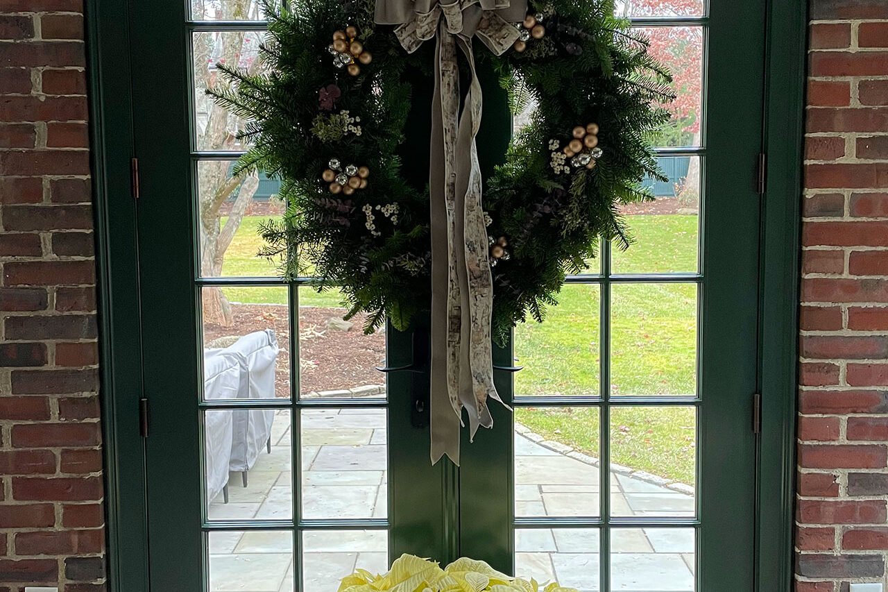 A lush evergreen wreath softens French doors, blending seamlessly with surrounding greenery for a classic New England feel.