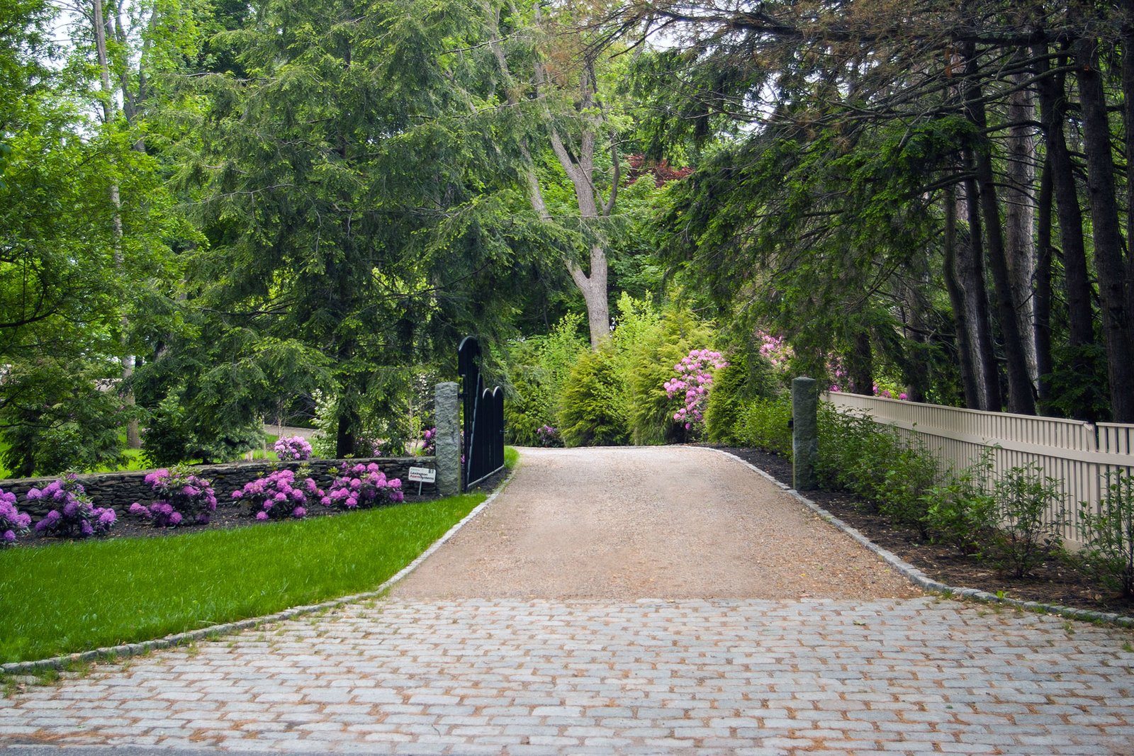 A cobblestone apron transitions into a gravel drive, with stone pillars marking entry and reinforcing a refined approach through a forested landscape.