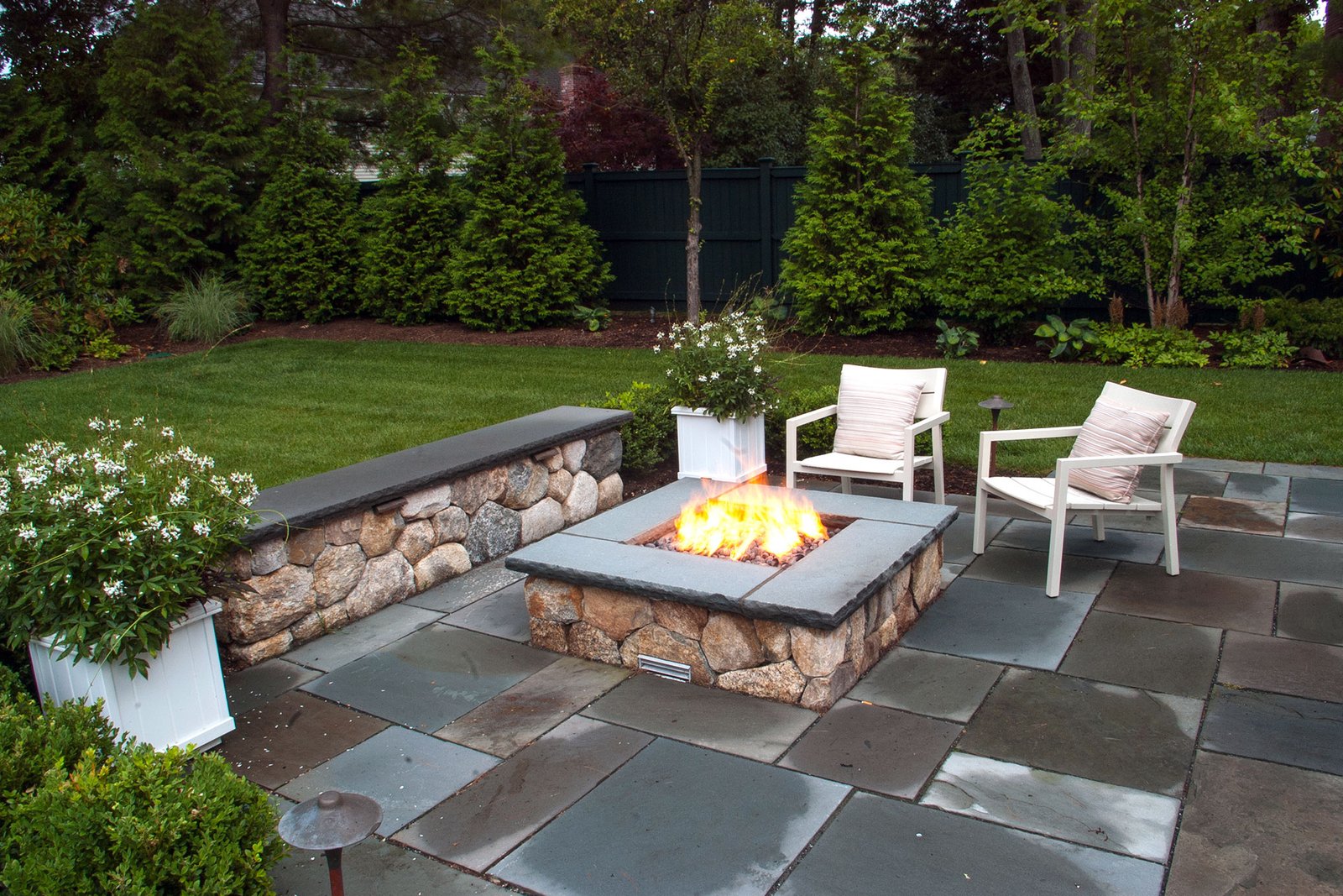 This natural stone fire pit with a seating wall in Weston, MA blends into the bluestone patio, providing warmth, safety, and a defined gathering space that feels both refined and relaxed.