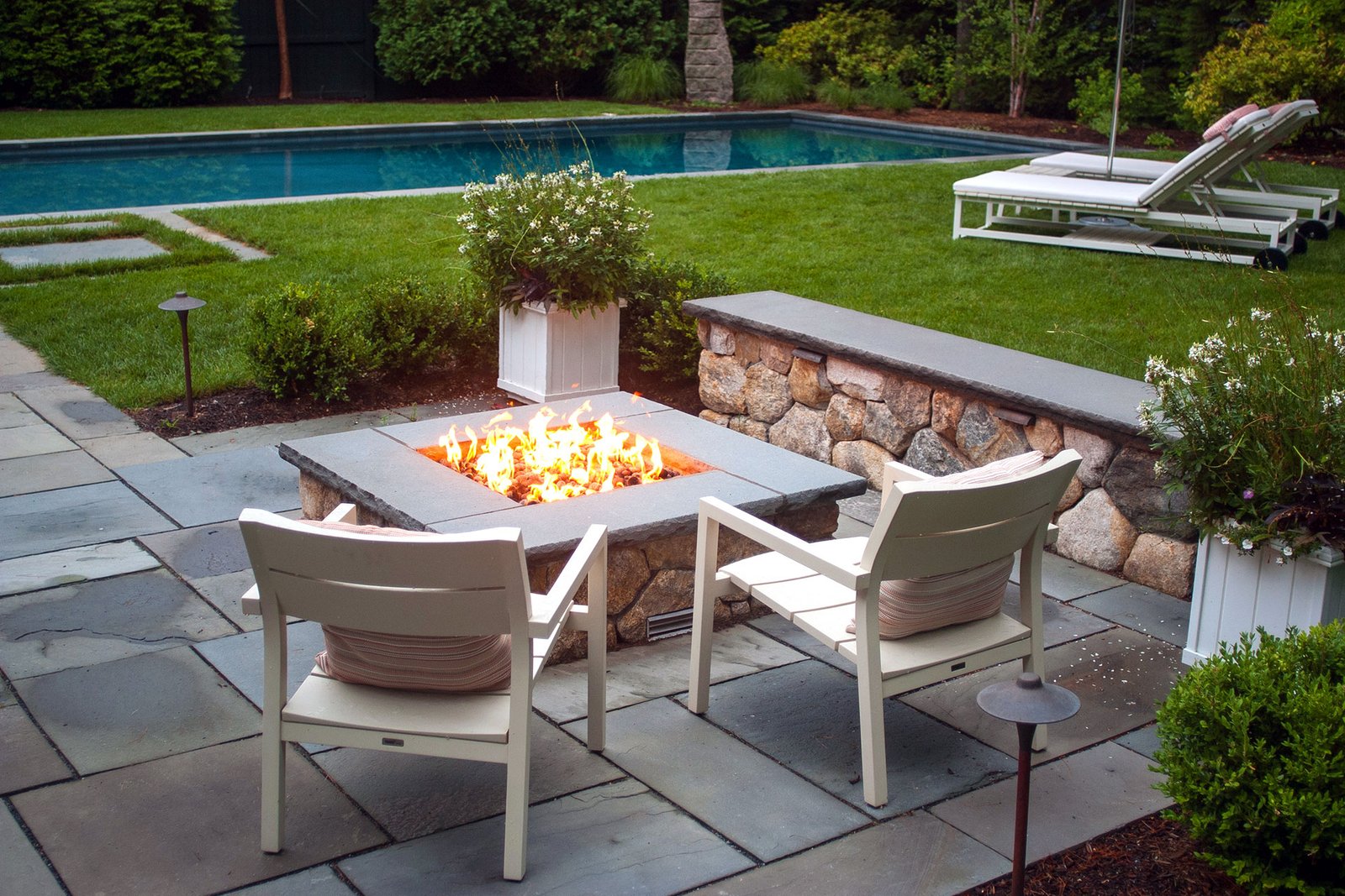 A square stone fire pit anchors this bluestone terrace beside the pool, offering clean lines, durable materials, and a central gathering point that enhances both day and evening use.