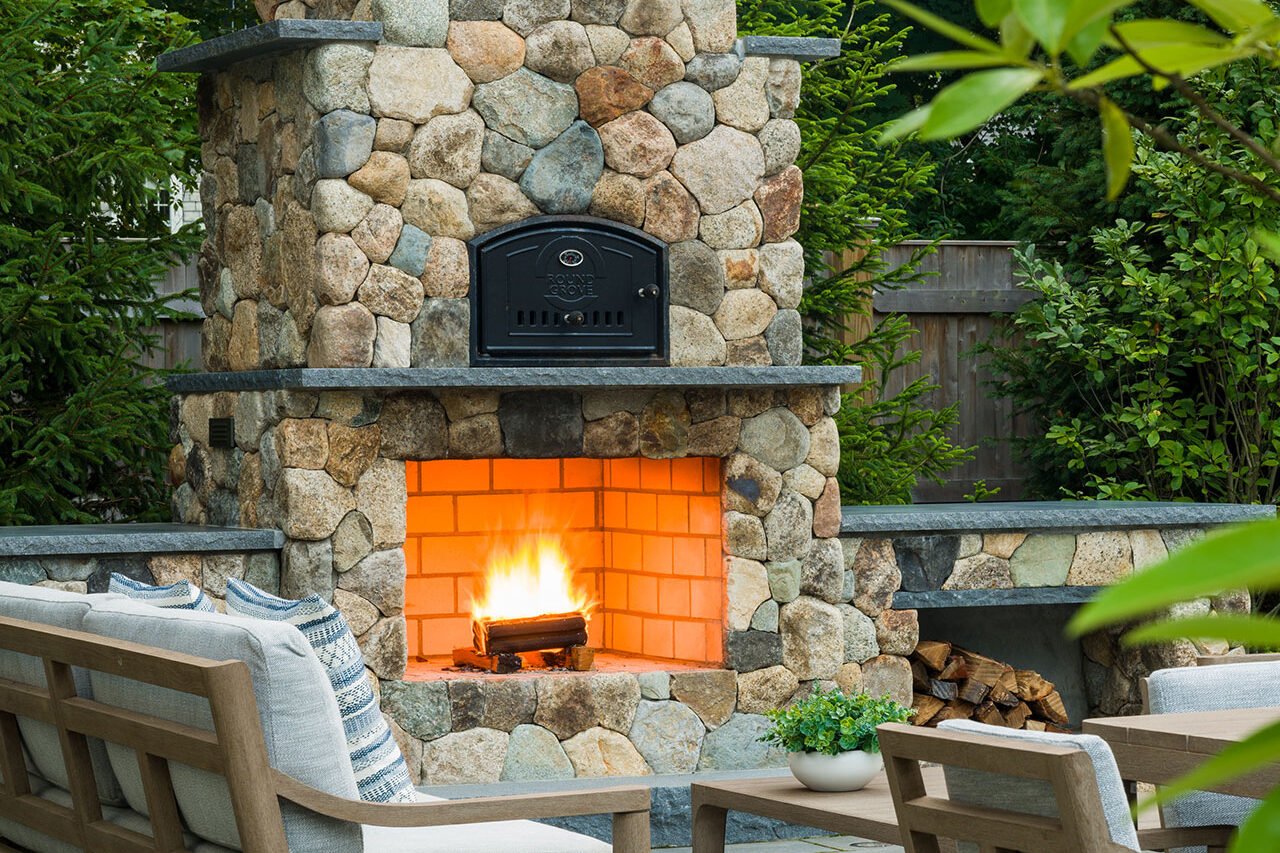 This tall natural stone outdoor fireplace features a wood-burning hearth that anchors the patio, offering radiant warmth, vertical presence, and a timeless focal point for evening entertaining.