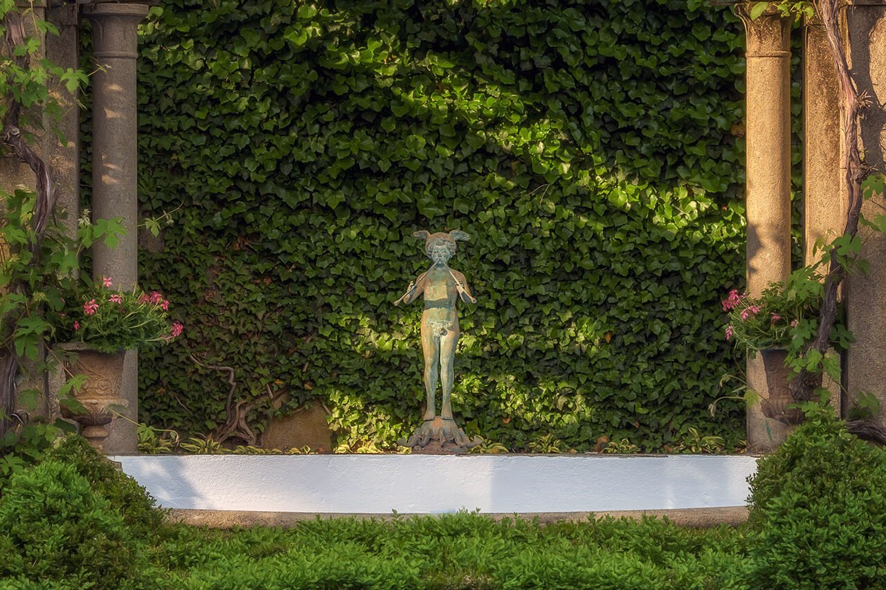 A classical figurative statue set against an ivy-covered wall and pergola creates a romantic focal point, blending sculpture, architecture, and lush greenery.
