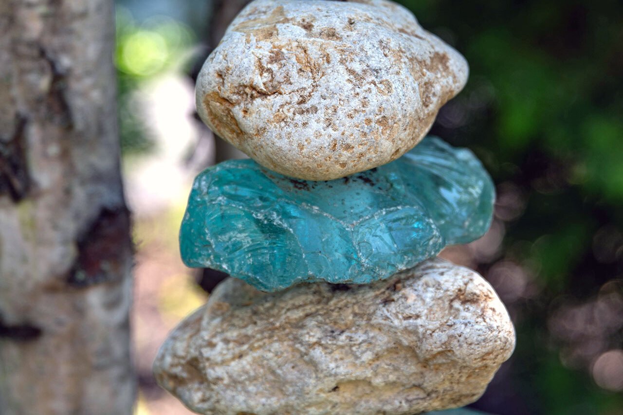 A stacked stone and glass cairn sculpture introduces vertical interest to the woodland garden, combining translucent blues with natural stone for a quiet, meditative focal point.