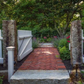 Lincoln, MA - A white garden gate framed by stone posts creates a graceful transition from walkway to garden, blending structure with softness.