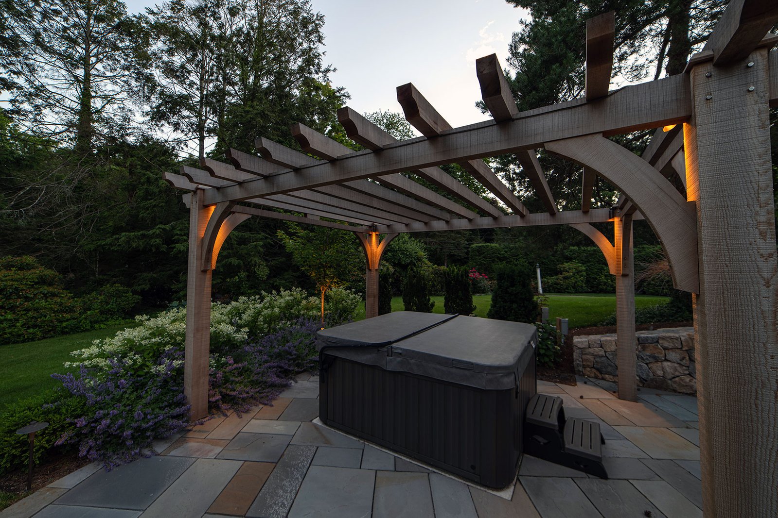 Cedar pergola with integrated lighting elevates a hot tub garden, extending use into the evening while adding privacy and structure.