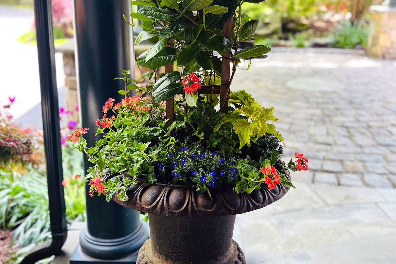 A rusted iron urn on a covered porch features mixed summer annuals, combining durability and warmth while reducing maintenance through sheltered placement.