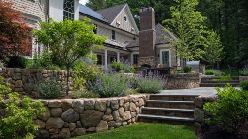 Tiered fieldstone walls paired with bluestone steps create a strong visual connection between the patio and surrounding planting, using layered stonework to manage grade while framing shrubs and low hedges that soften transitions between outdoor rooms.