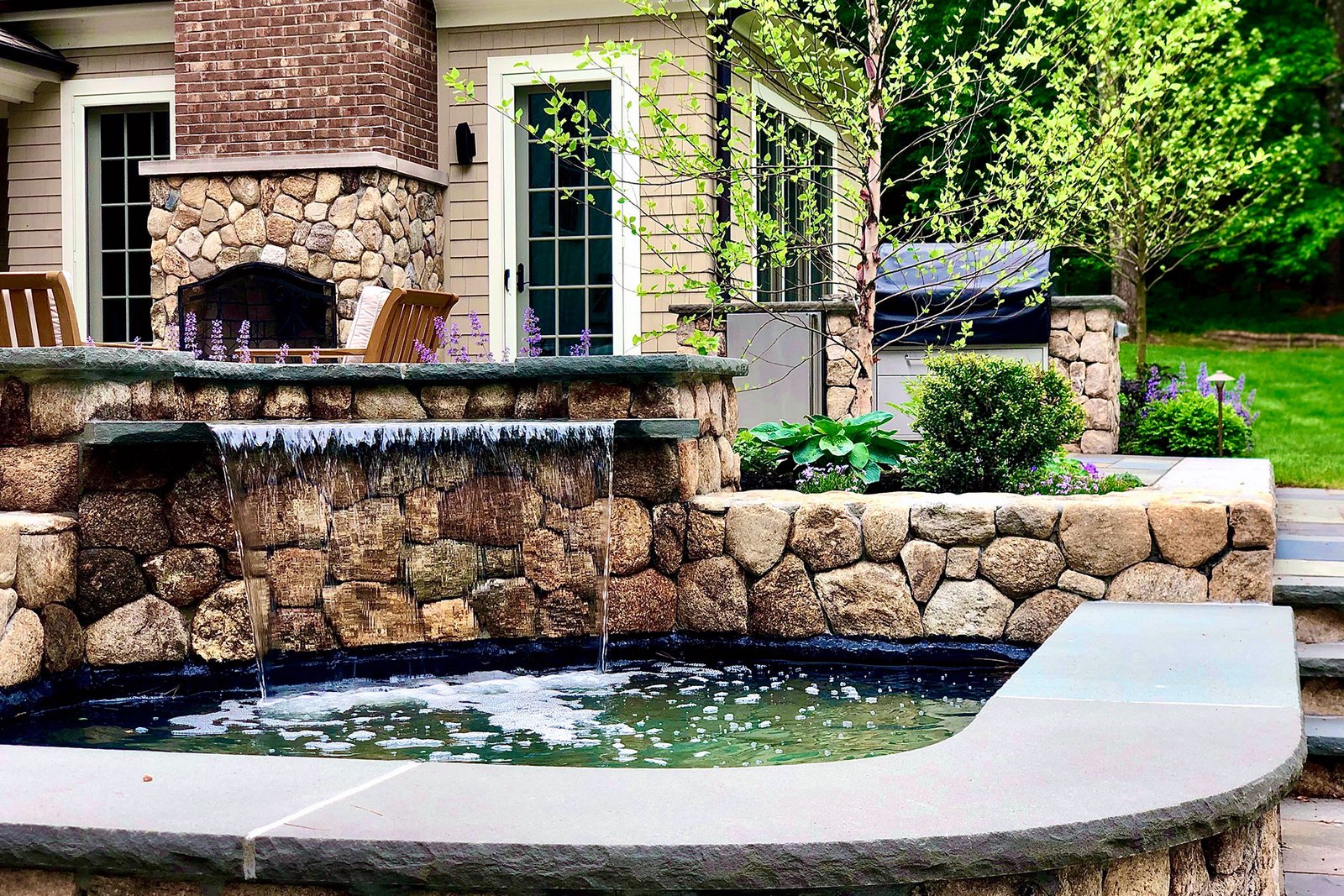 A hand-built stone wall waterfall enhances this patio with gentle sound, visual texture, and a seamless connection between hardscape and surrounding landscape.