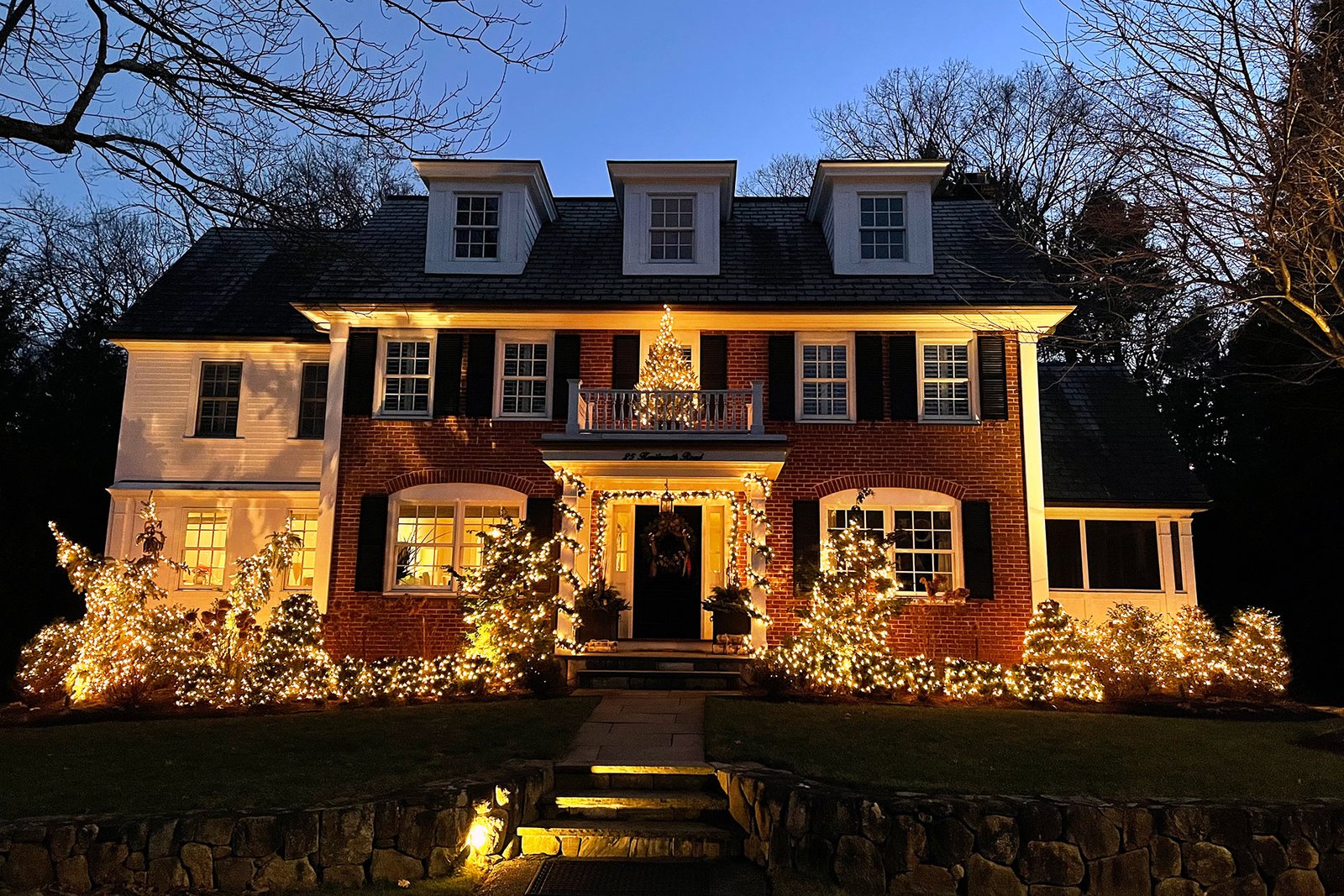 Colonial-style home illuminated at night with warm white holiday lights and seasonal landscape decorations.