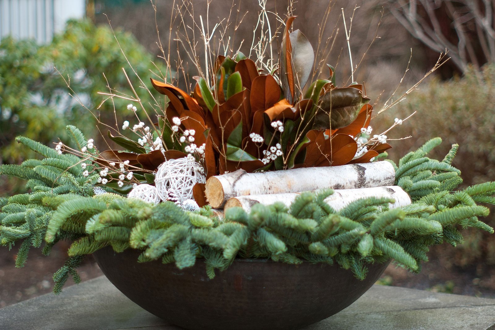 Evergreen bowl planter arranged with birch logs, magnolia leaves, pinecones, and seasonal greenery for an elegant holiday display.